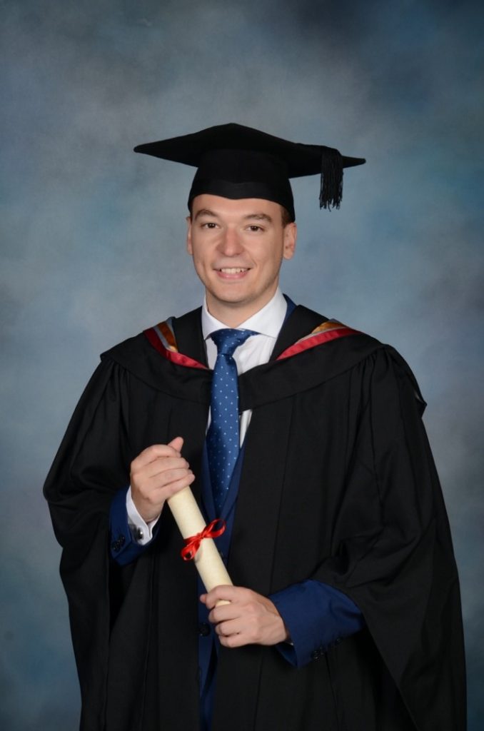 Alvaro Lopez Pellicer graduating from Lancaster University. He is holding a scroll in his hands and is dressed in Lancaster University robes (black with a gold, grey and red hood, with a black mortar board).