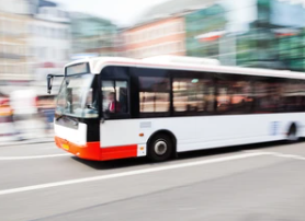 A white bus driving in an urban environment. The bus is shown as being rather blurry, indicating that it must be travelling at speed