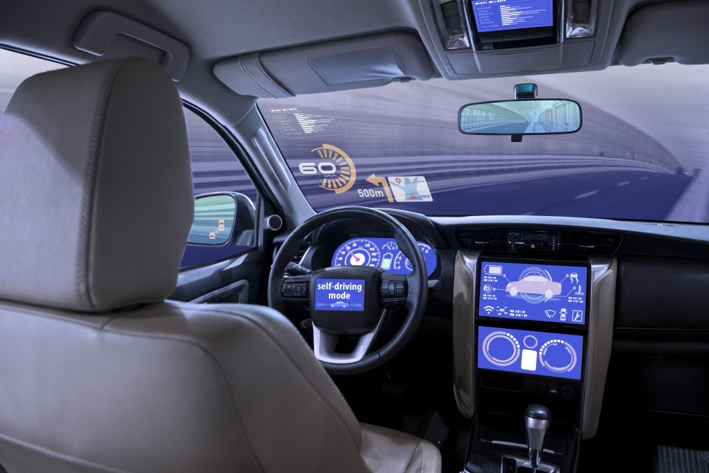 Driver's seat and dashboard of a left-hand drive autonomous car. There is nobody sitting in the drivers seat and the computerised displays on the dashboard and steering wheel indicate that the car is in self-drive mode.