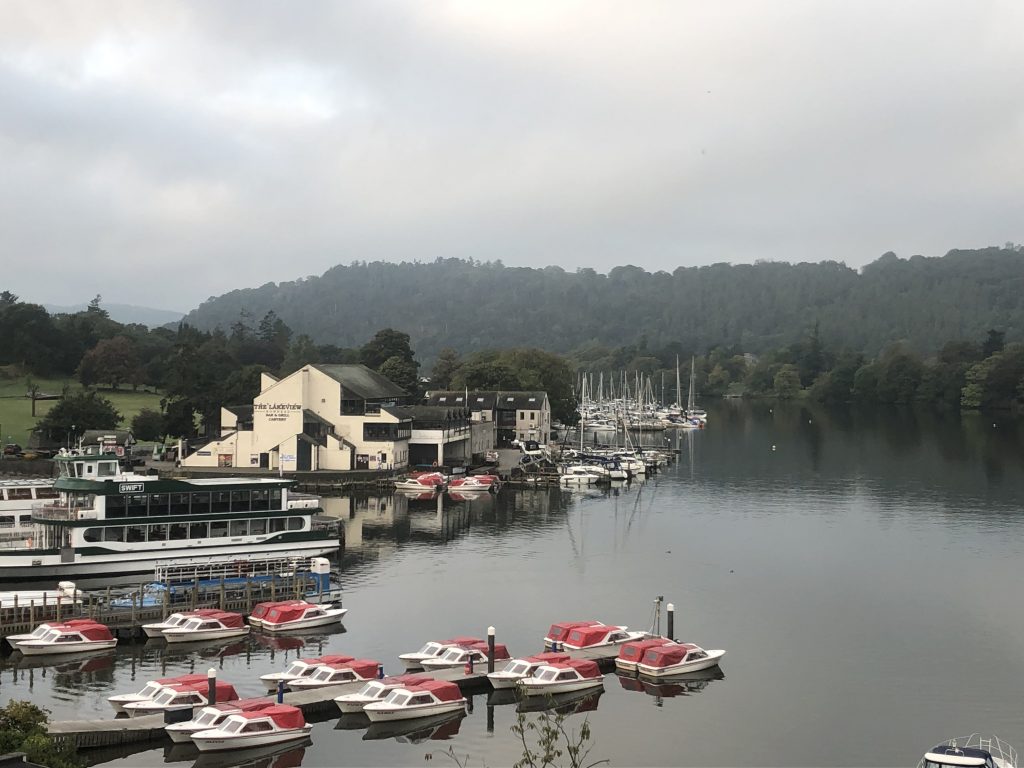 A rather cloudy Lake Windermere. A selection of boats can be seen at Bowness Pier, with the Lakeview Restaurant behind them. In the background, there are the hills and tress of the surrounding countryside.
