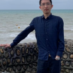 Yi Li standing by a wall with the sea behind him